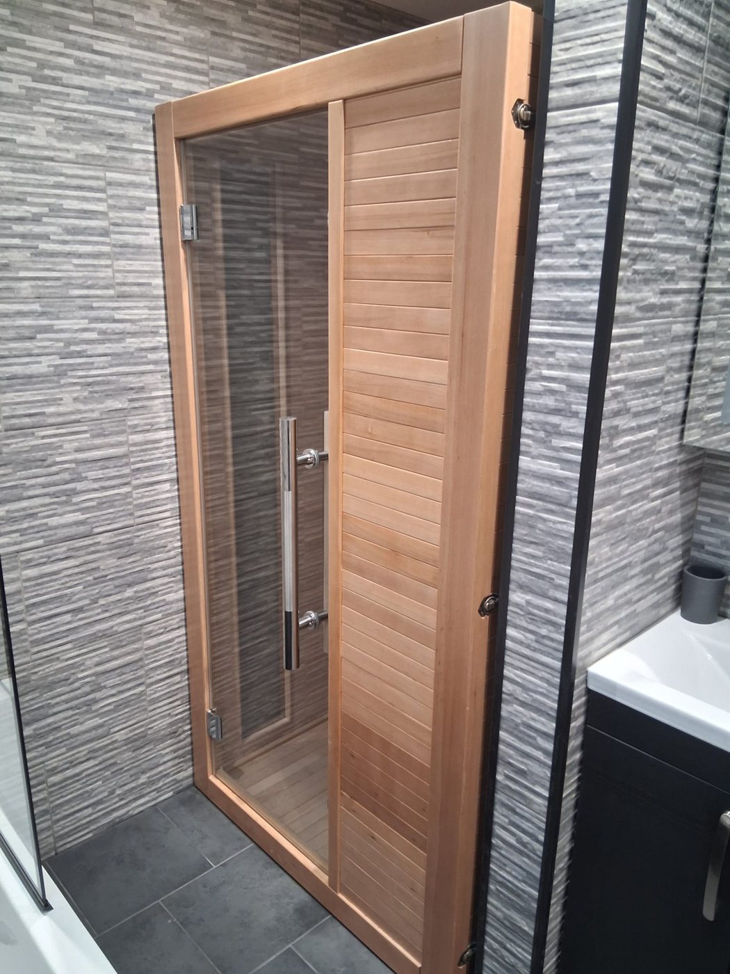 Timber sauna cabin with a glazed door and slatted panel, set into a bathroom tiled in grey split-face effect tiles