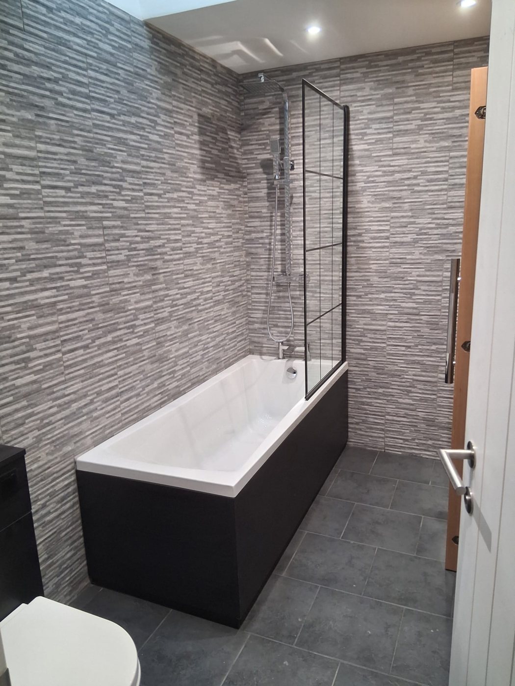 White bath with a dark side panel and a black grid shower screen against full-height grey wall tiles